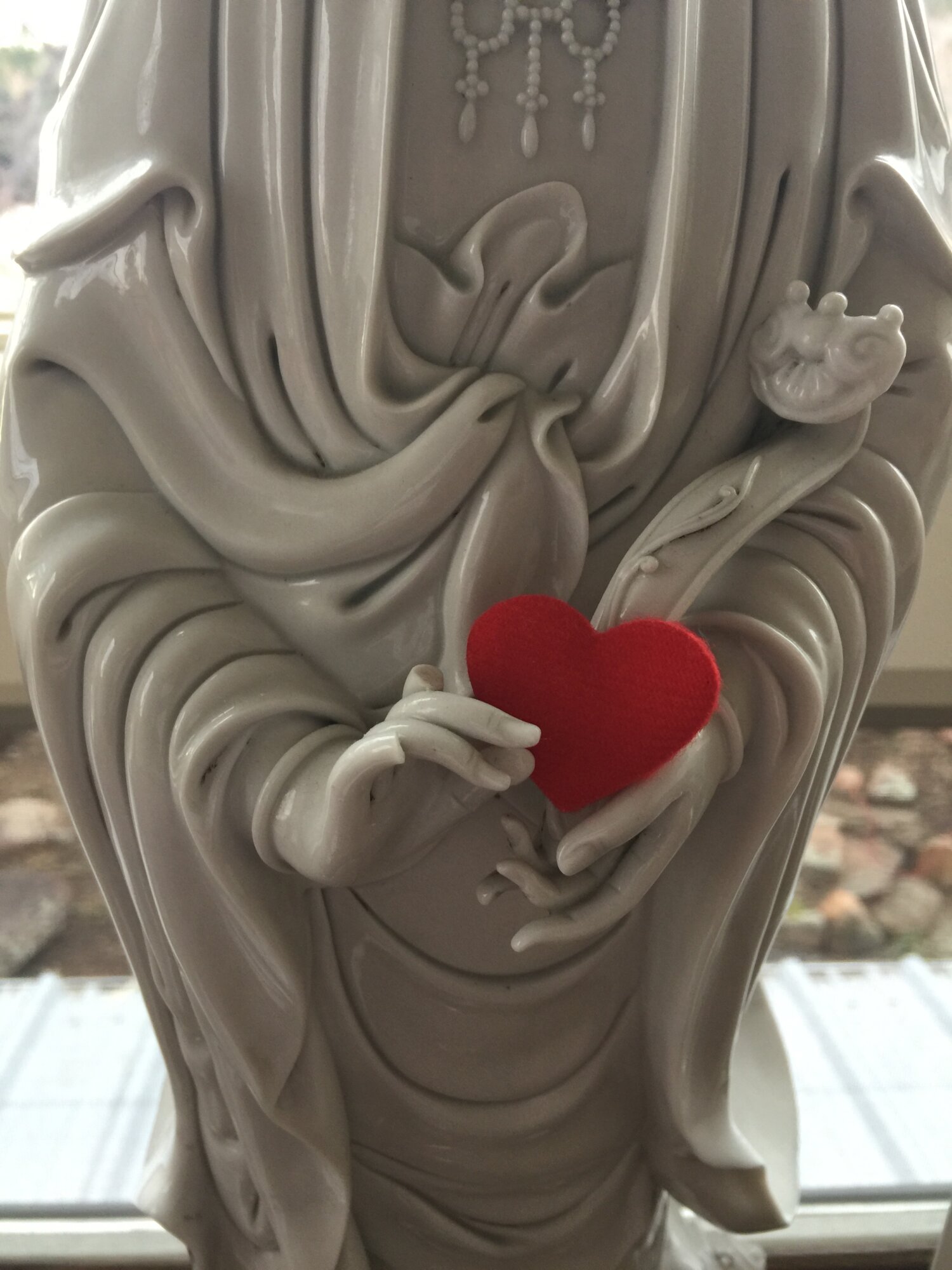 A partial image of a porcelain statue of Kwan Yin with a small red heart place in its hands.